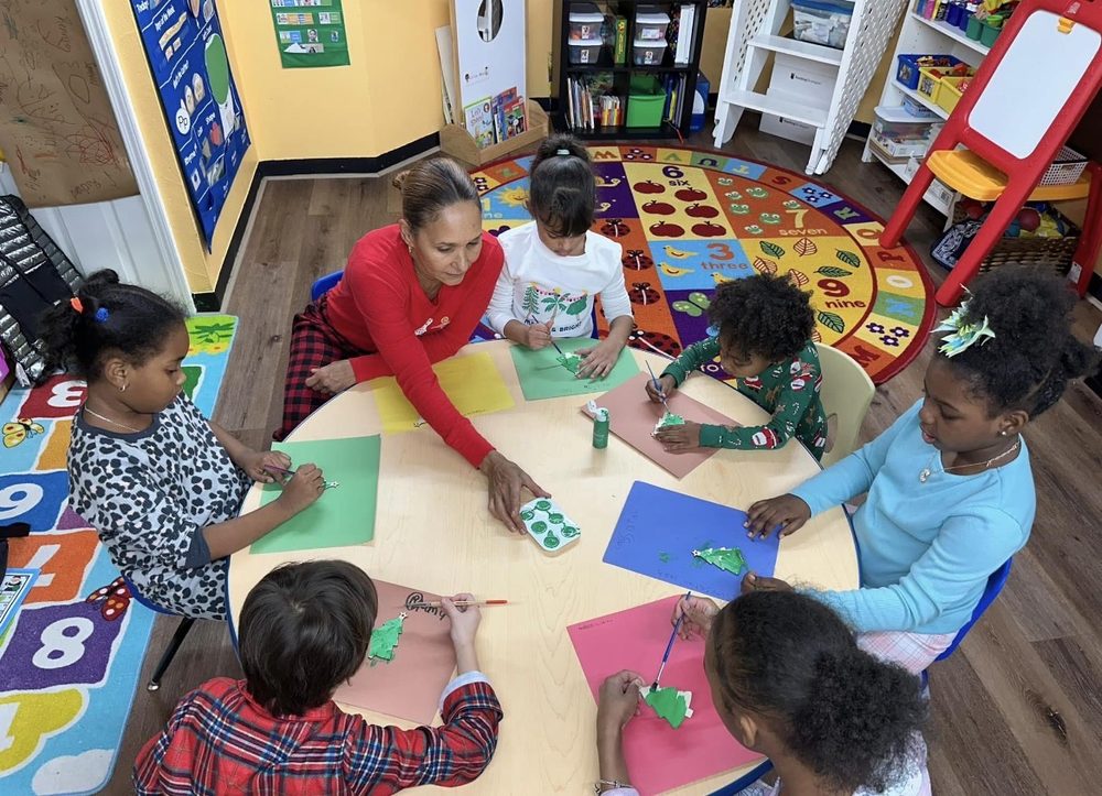Teacher helping a group of children paint holiday tree crafts at a classroom table
